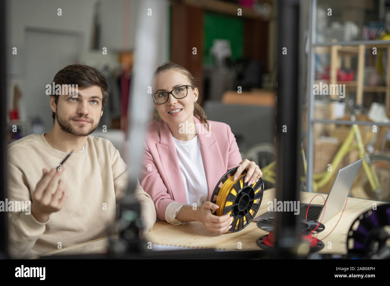 Two young creative designers looking at 3d printer while discussing its characteristics and model by workplace Stock Photo