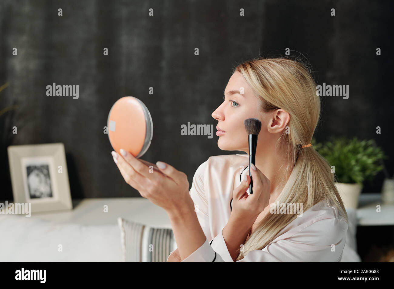 Pretty blond woman applying powder on face with brush while looking in mirror Stock Photo