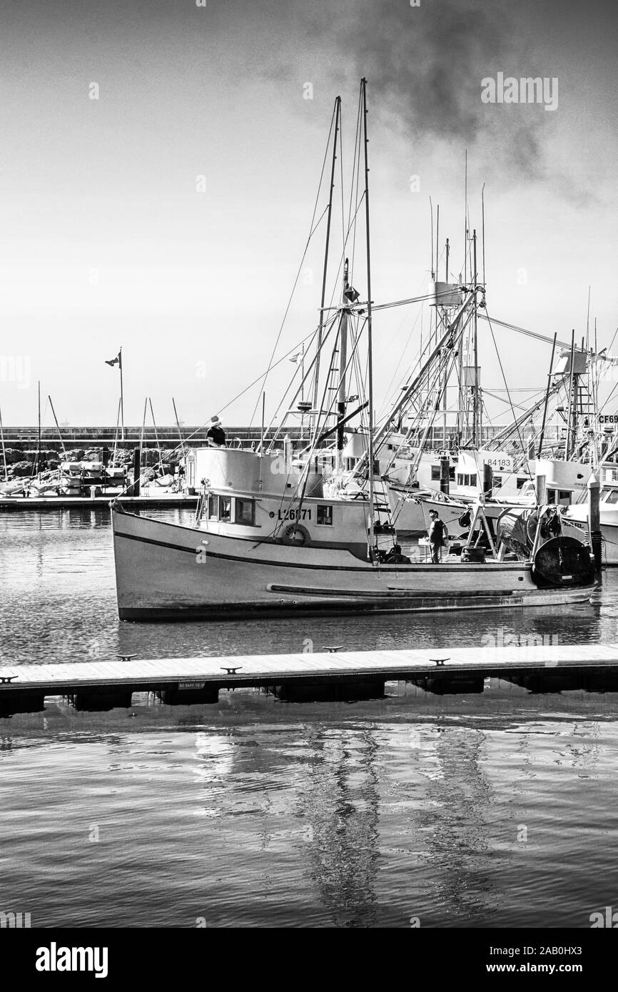An aged commercial fishing boat is launching from the docks of the Marina in the Santa Barbara Harbor, in Southern California, in black and white Stock Photo