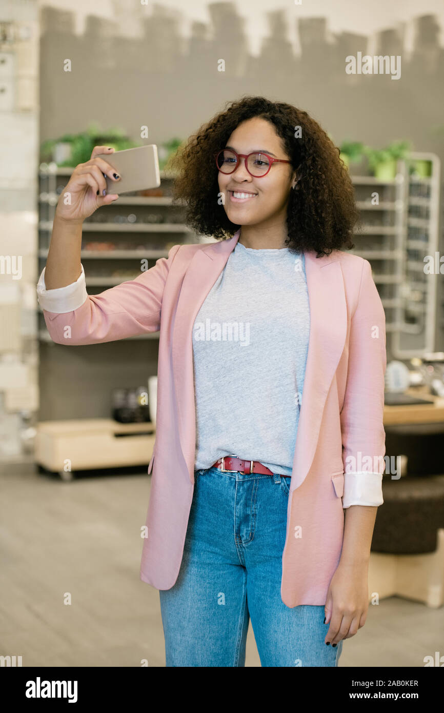 Happy mixed-race girl in smart casual making selfie while trying on new eyewear Stock Photo