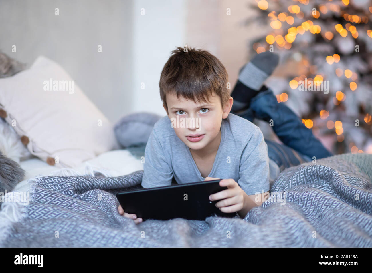 serious boy holding tablet in hands. child plays computer games on tablet. boy lies on bed opposite Christmas tree before Christmas. Black Friday and Stock Photo