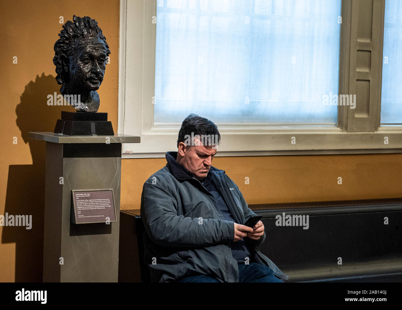 Man sitting on bench using mobile phone with sculpture of Einstein on plinth. Victoria and Albert Museum, South Kensington, London, England, UK. Stock Photo