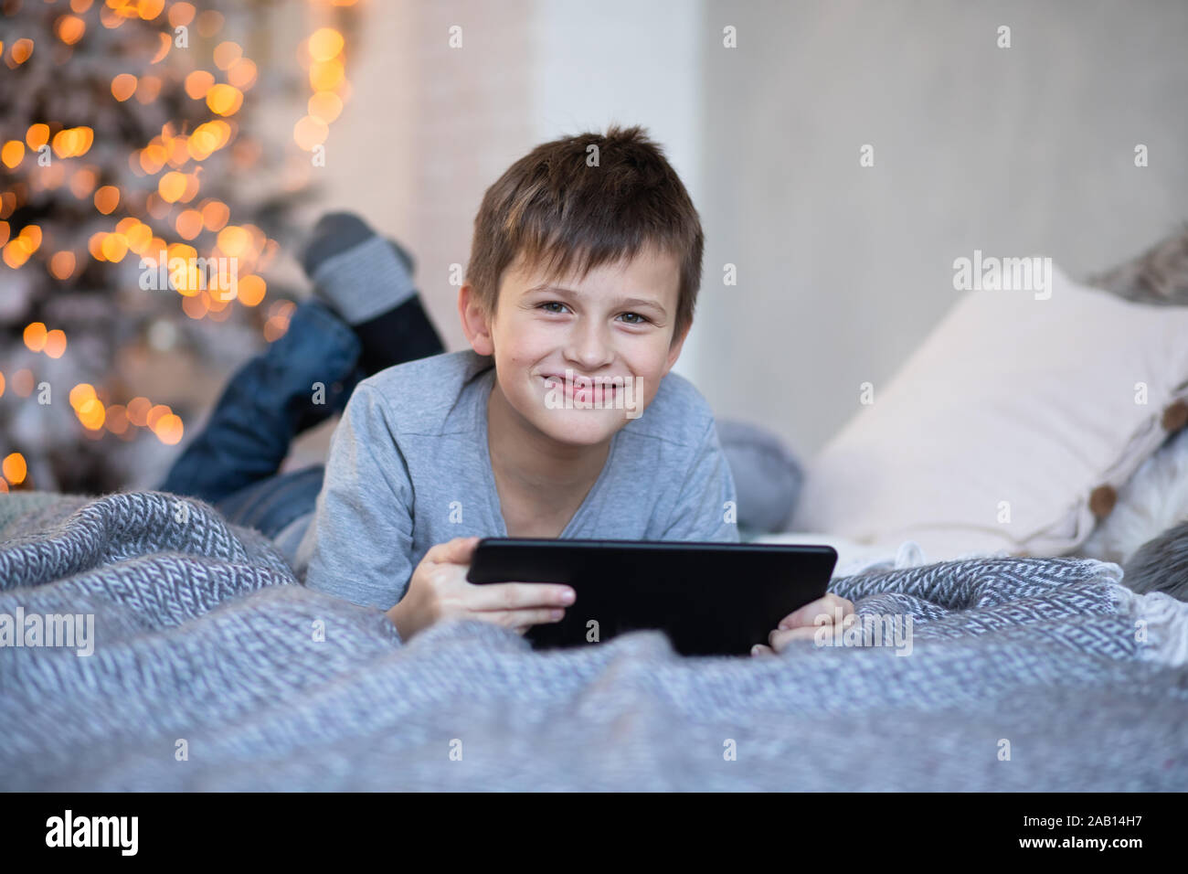 Happy handsome boy with a tablet in hands. child plays computer games on tablet. boy lies on bed opposite Christmas tree before Christmas. Black Frida Stock Photo