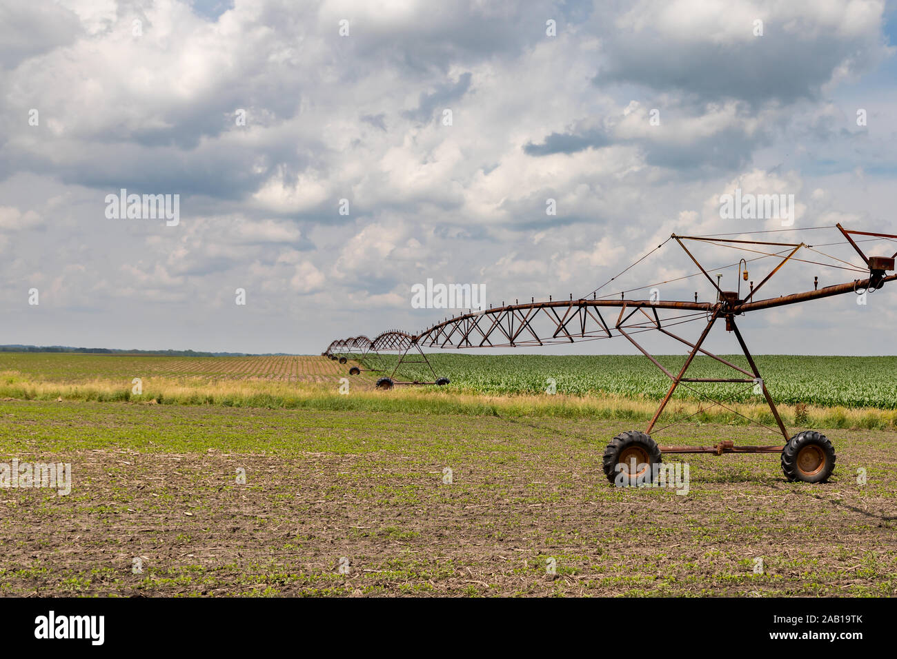 Rusty, old center pivot irrigation system stretching over rolling hills between growing soybean and corn farm fields. Sunny summer day with clouds Stock Photo