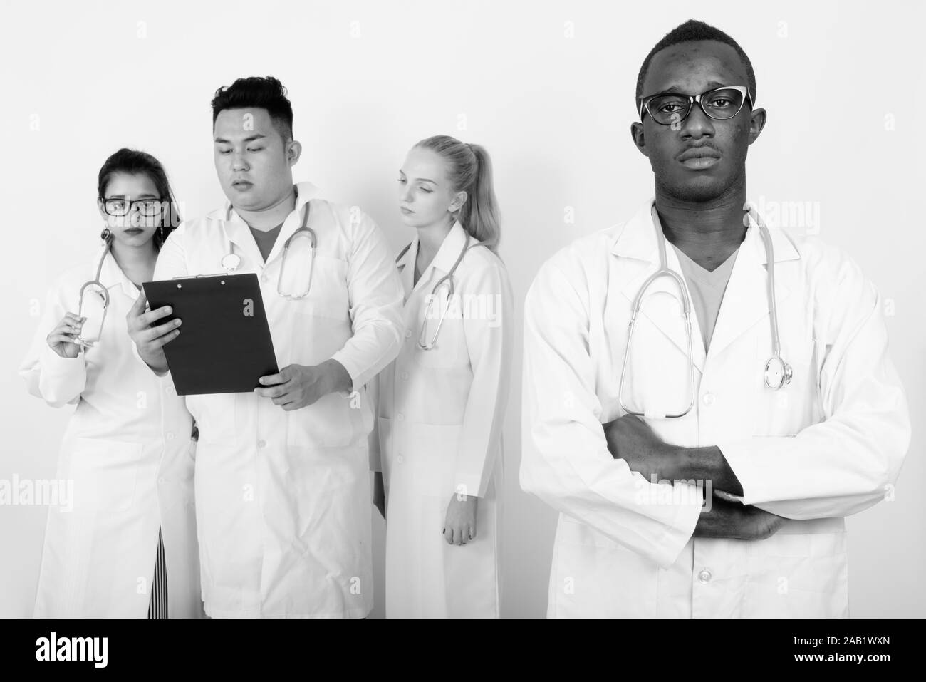 Studio shot of young African man doctor with arms crossed and diverse group of multi ethnic doctors reading on clipboard together Stock Photo