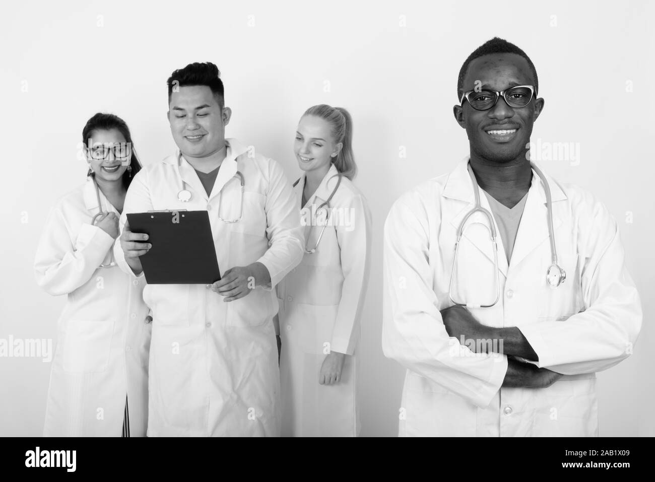 Studio shot of happy young African man doctor with arms crossed and diverse group of multi ethnic doctors smiling while reading on clipboard together Stock Photo