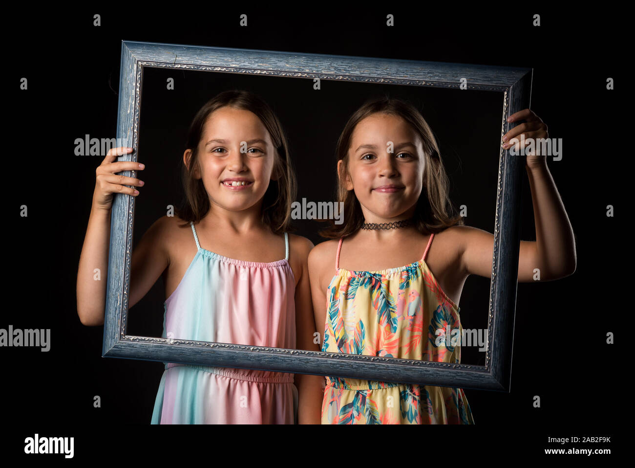 Identical twin girls are making happy expressions with picture frame. Children, sisters, girls posing in studio with picture frame, making different f Stock Photo