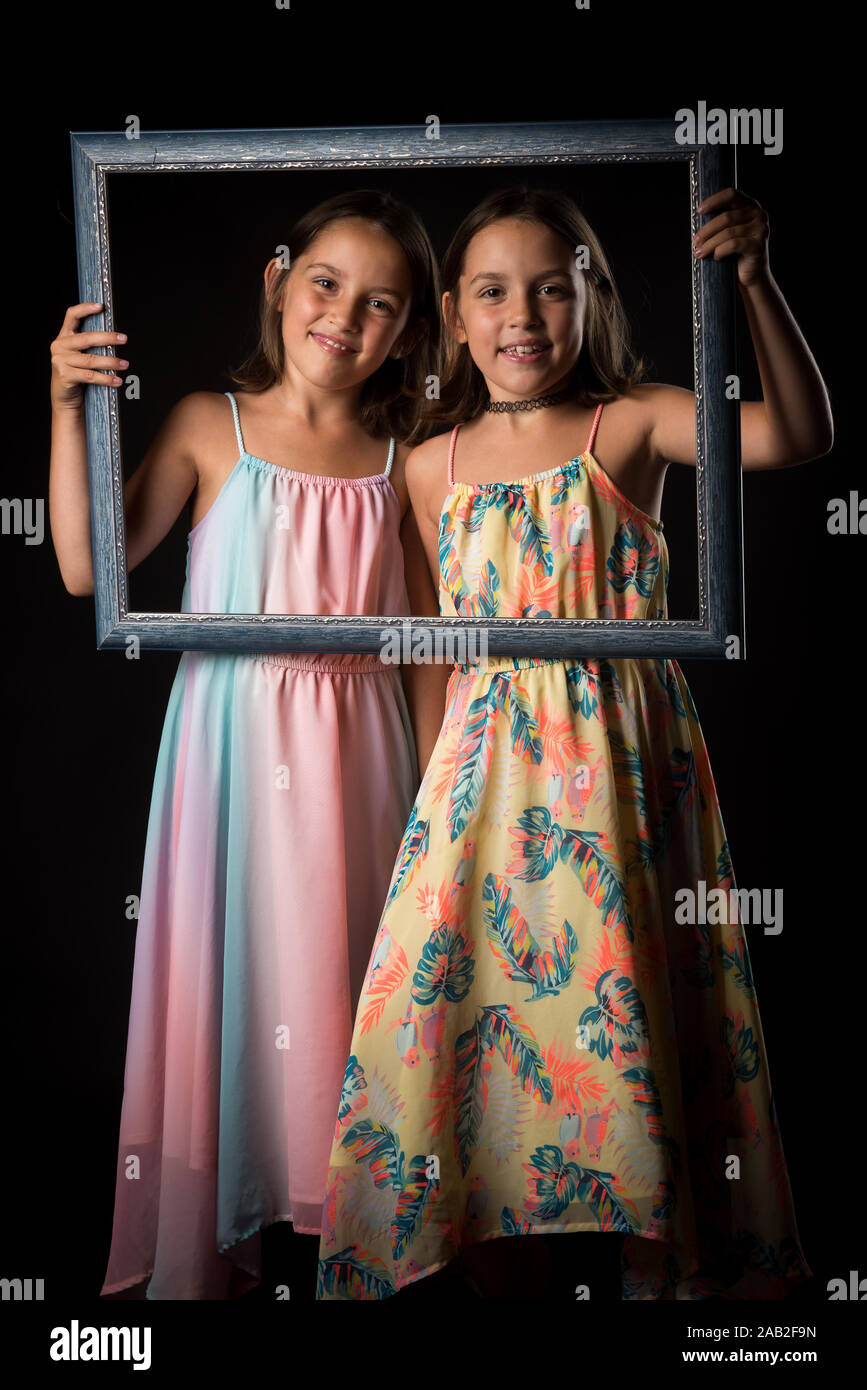 Identical twin girls are making happy expressions with picture frame. Children, sisters, girls posing in studio with picture frame, making different f Stock Photo