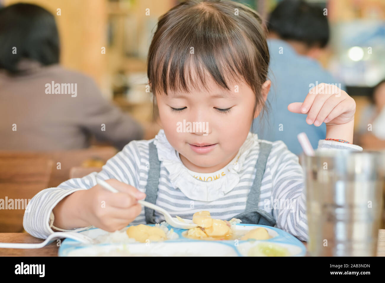 Little asian child girl use spoon to scoop food on the table to dine. while having lunch on table in restaurant Stock Photo