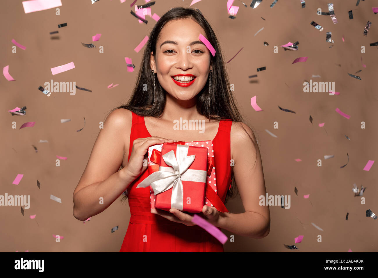 Freestyle. Young girl in dress standing in falling confetti isolated on brown with presents laughing happy Stock Photo