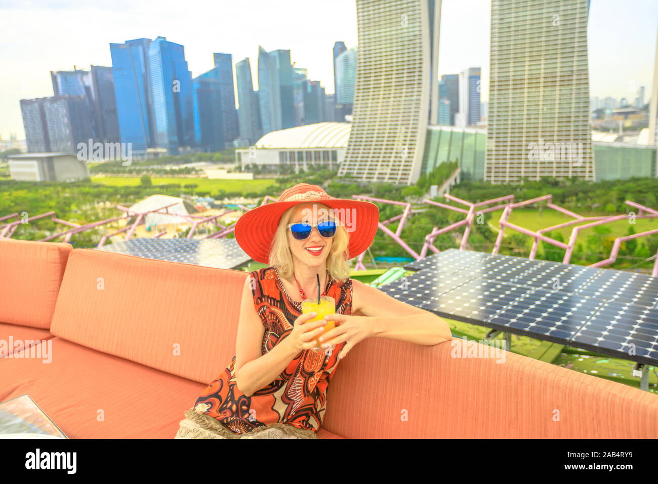 Happy tourist enjoying a drink on holiday in Singapore, Southeast Asia. Lifestyle caucasian woman with red hat drinking aperitif at rooftop. Aerial Stock Photo