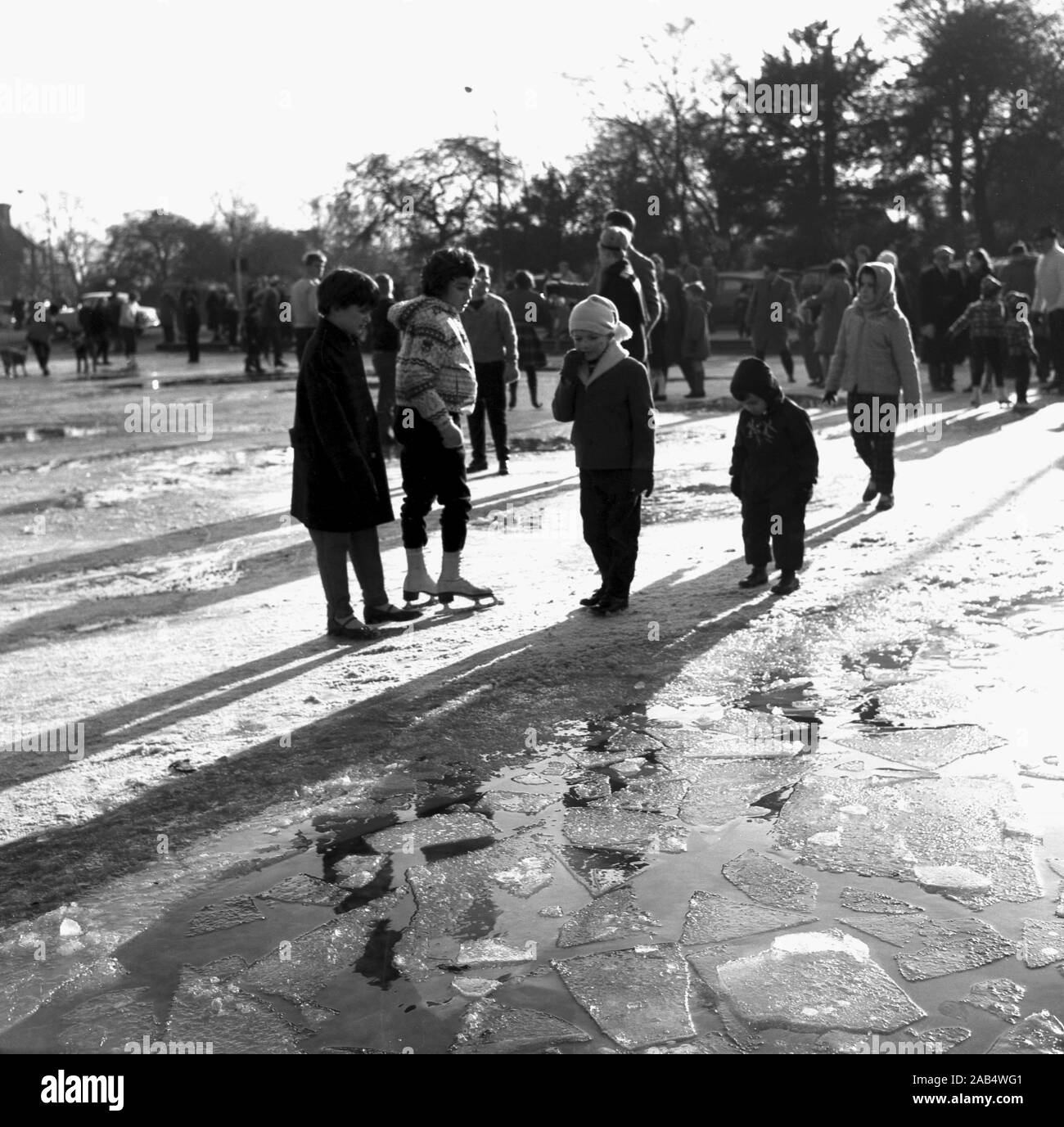 1960s, historical, wintertime and several children, some wearing ice skates, standing outside on a path by on a part frozen, icy lake, England, UK. Stock Photo