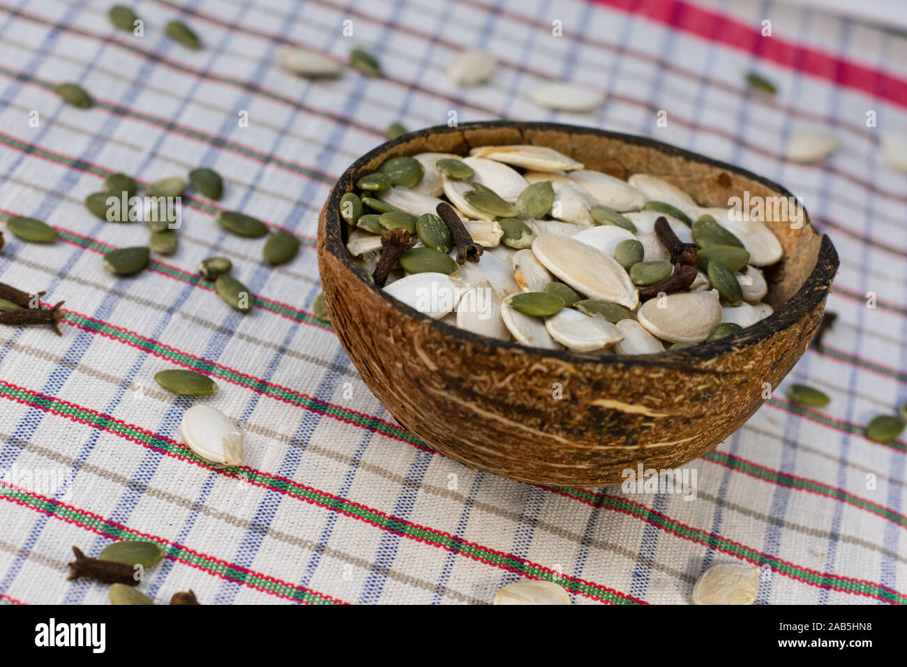Coconut bowl full of peeled and unpeeled pumpkin seeds in white background with scattered pumpkin seeds and clove spice. Stock Photo