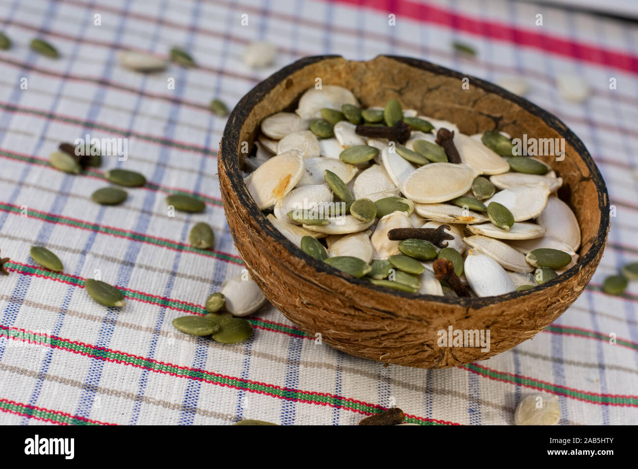 Coconut bowl full of peeled and unpeeled pumpkin seeds in white background with scattered pumpkin seeds and clove spice. Stock Photo