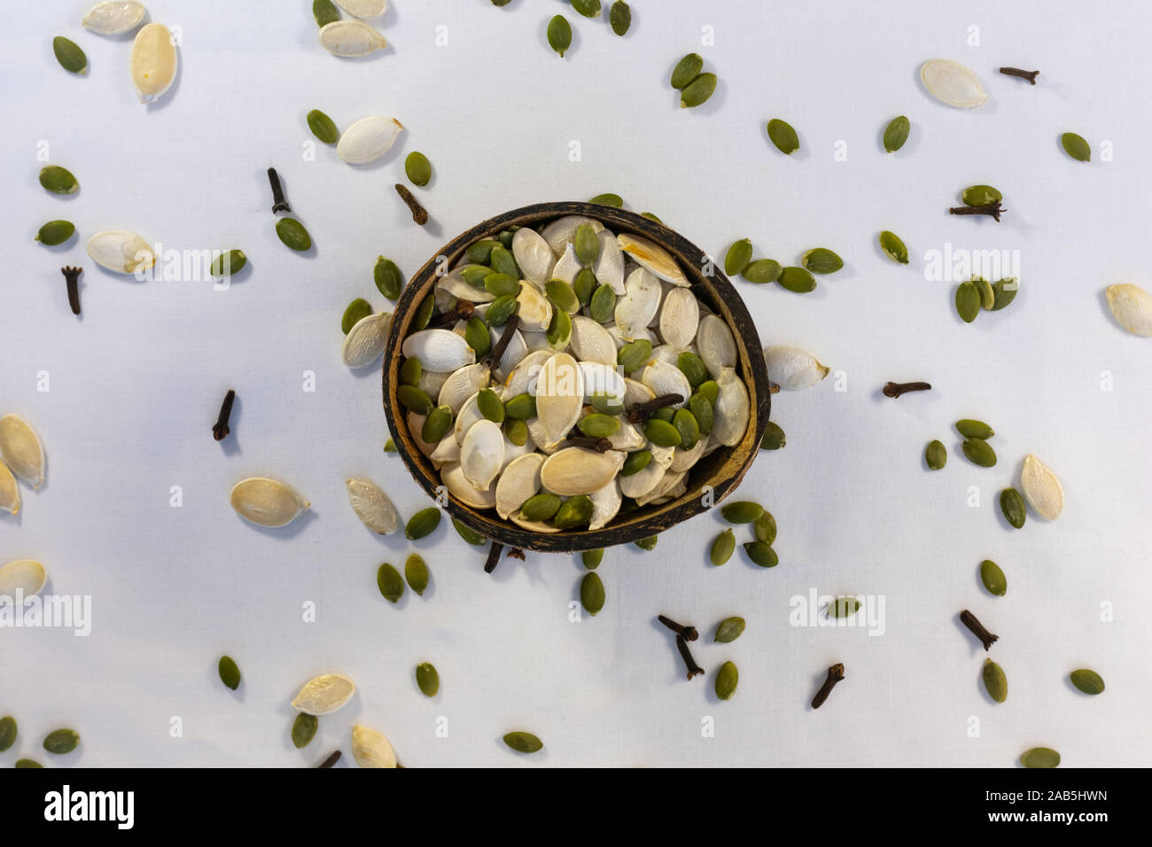 Coconut bowl full of peeled and unpeeled pumpkin seeds in white background with scattered pumpkin seeds and clove spice. Stock Photo