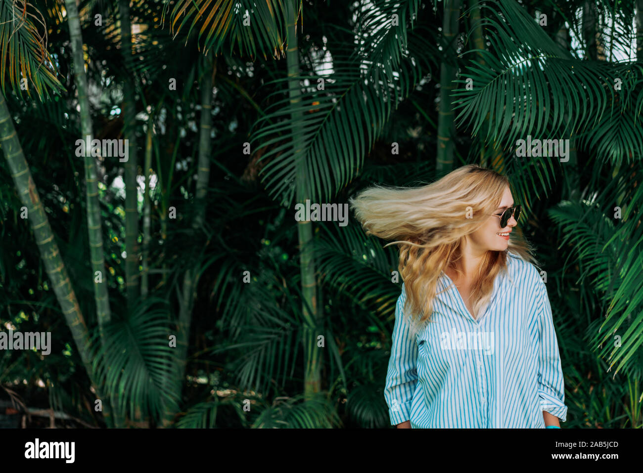 Portrait of beautiful of traveler woman with sunglasses smiling behind palm trees. Sunlight. Concept travel, vacation, dream. Flying hairstyle Stock Photo