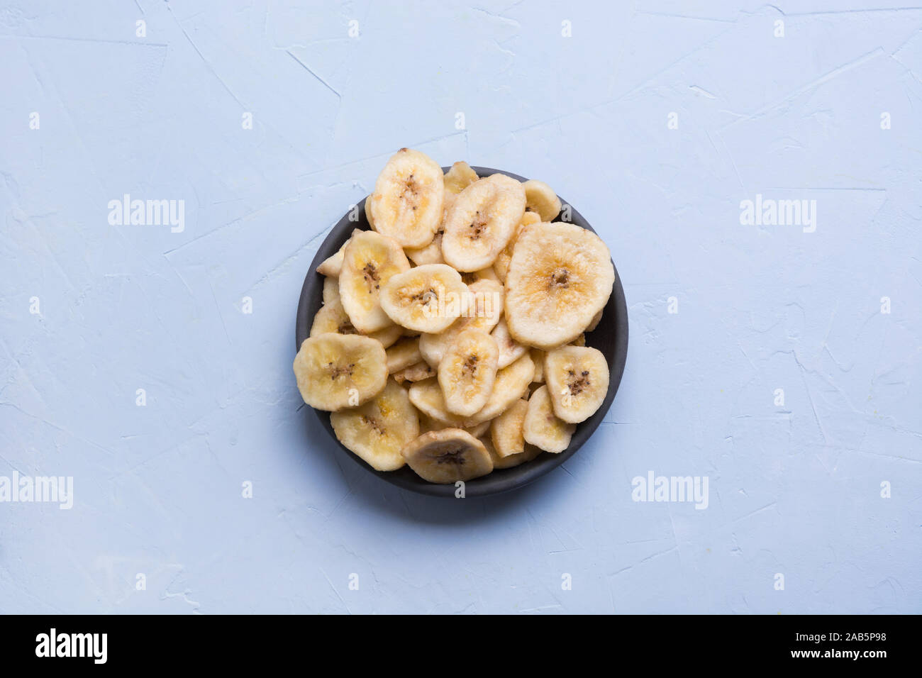 Dehydrated, fresh and dried banana chips in black bowl on light background. View from above