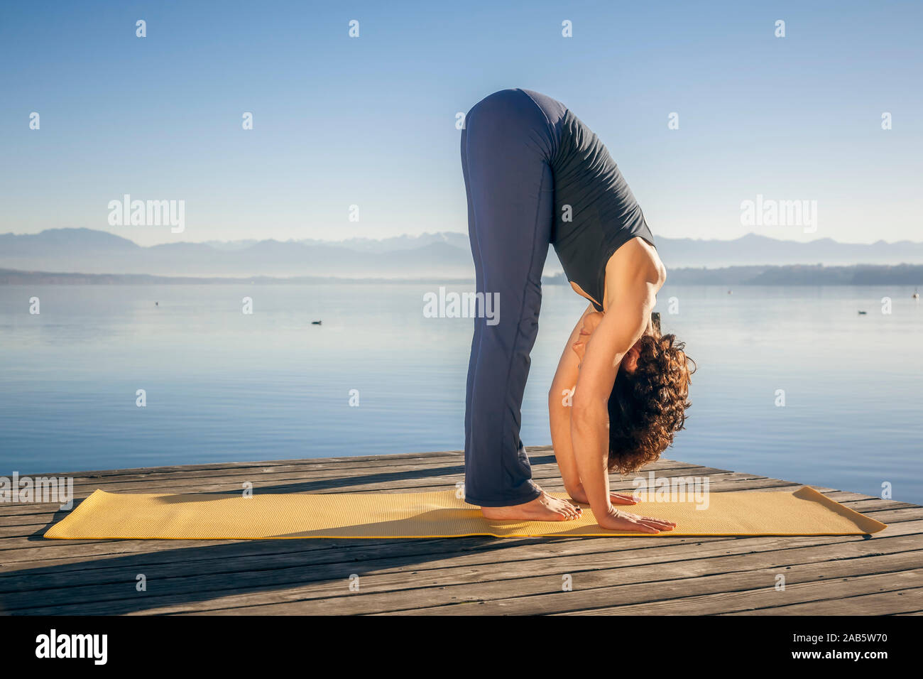 Eine junge Frau, die Yoga-Uebungen am Seeufer durchfuehrt (Uttanasana) Stock Photo