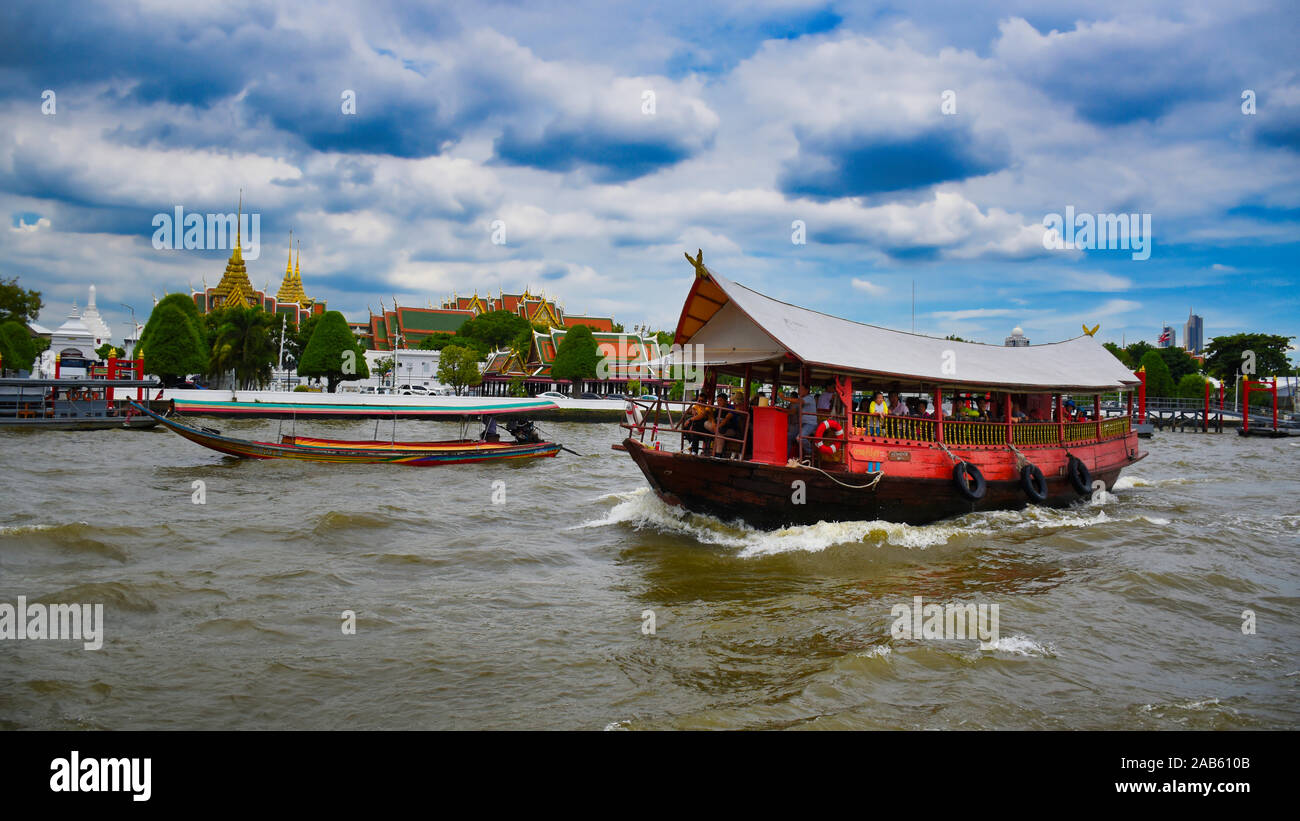 Bangkok, Thailand .11.24.2019: A red wooden traditional boat with tourist and a long tailed boat is sailing the waves of the Chao Phraya River in fron Stock Photo