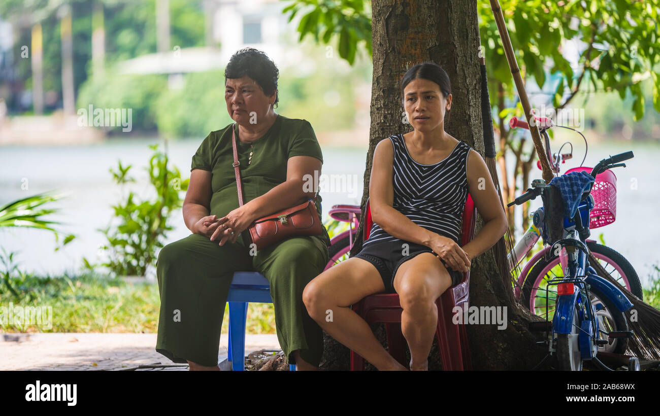 Hanoi, Vietnam - 11th October 2019: Two Asian women sit in the shade under a tree on plastic chairs during a heat wave in Asia Stock Photo