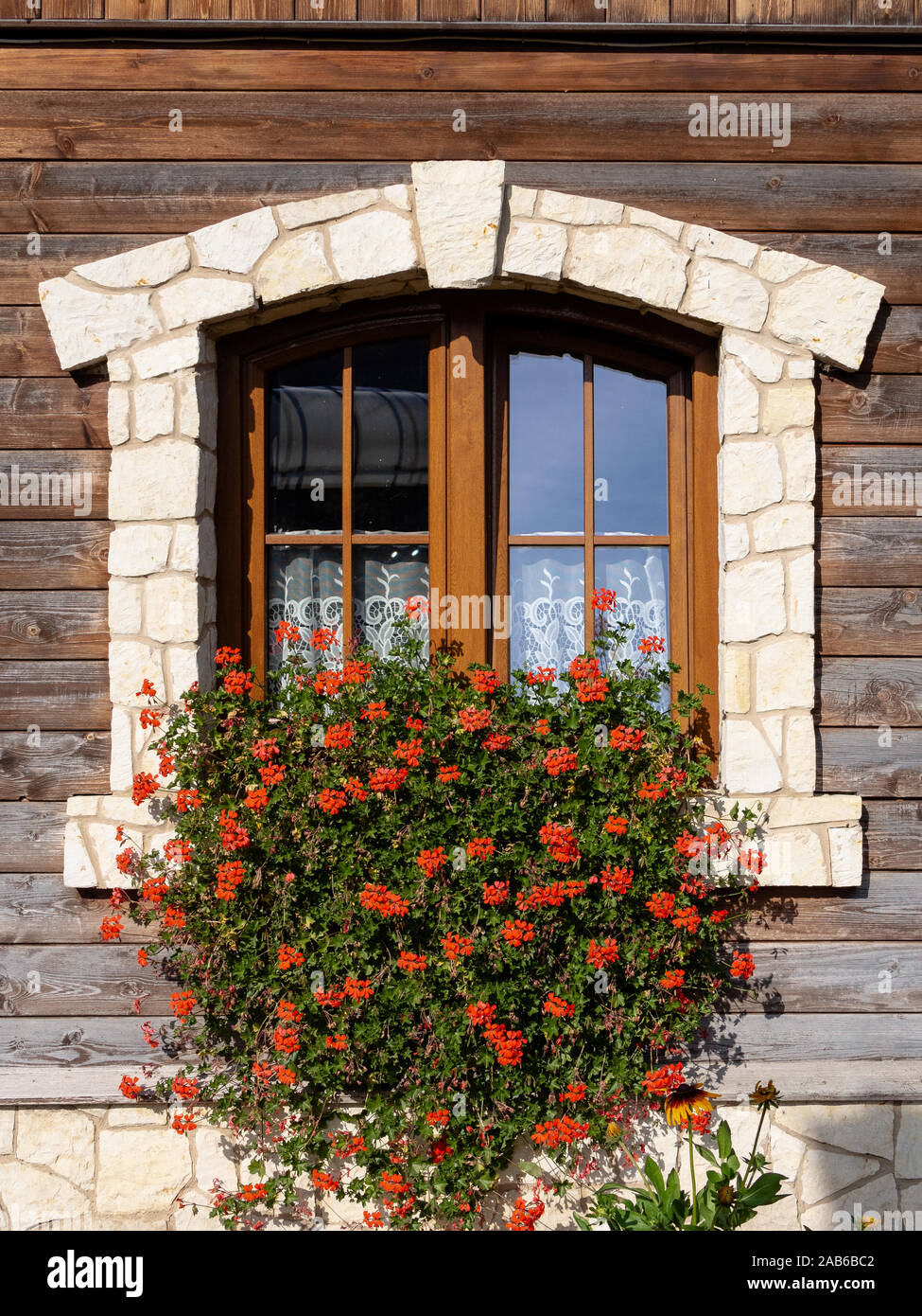 Window of a rural wooden house. A beautiful, flowered, stone-framed window in the wooden wall of the house. Bright, warm colors. Daylight. Stock Photo