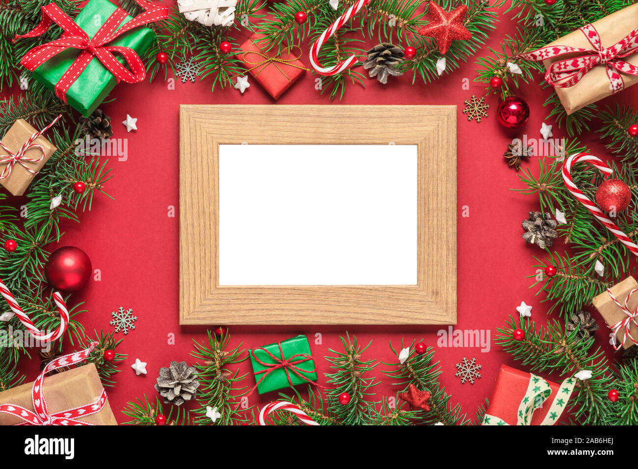 Christmas photo card in frame made of fir tree branches, gift boxes, candy, holiday decorations on red background. mock up. flat lay. top view with co Stock Photo