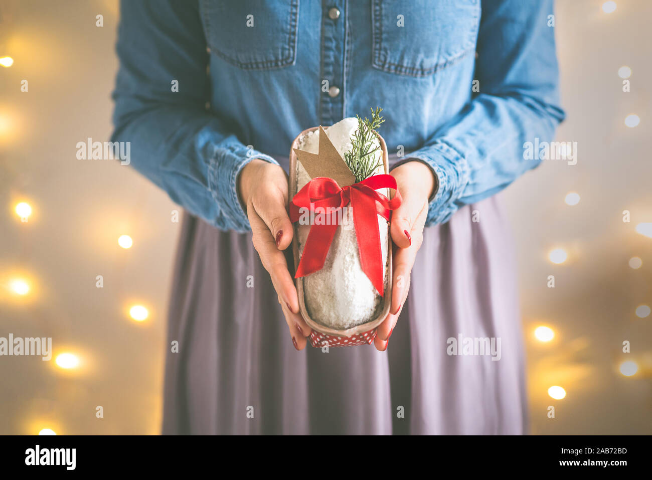 Close up of woman's hands holding homemade stollen cake wrapped as a gift in front of the christmas lights. Selective focus. Stock Photo