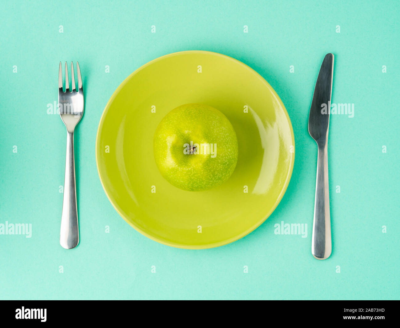 Ripe raw juicy green Apple on a yellow plate, fork, knife on a bright turquoise blue colour table, top view. Diet as a lifestyle. Stock Photo