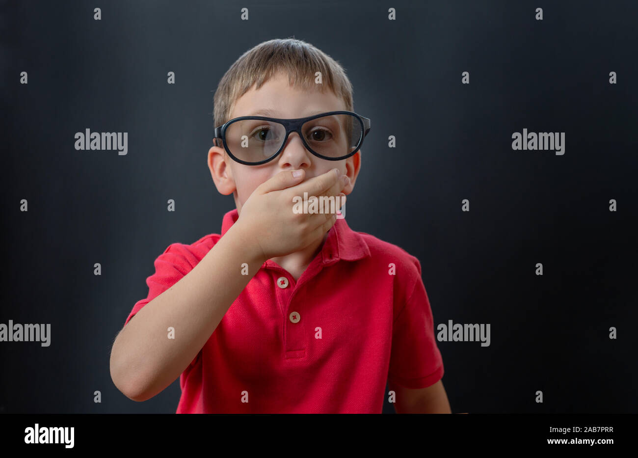 The boy is anxiously watching a movie with 3d glasses. He holds his hand to his mouth and looks surprised. Stock Photo