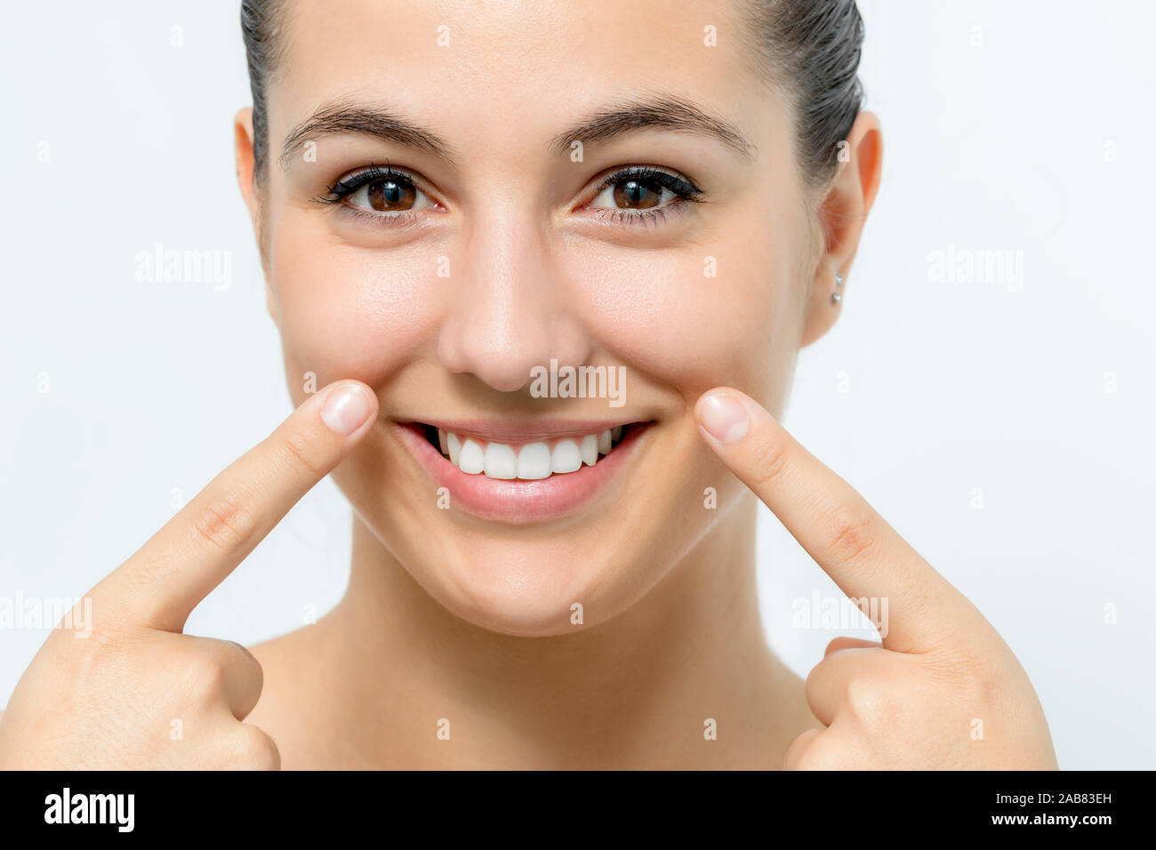Extreme close up portrait of cute young woman pointing at wrinkles below cheek. Girl with charming smile and healthy white teeth isolated on white bac Stock Photo