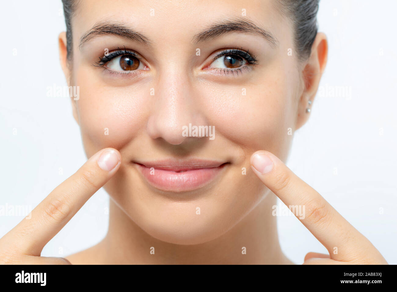 Extreme close up portrait of cute young woman pointing at wrinkles below cheek. Girl with charming smile looking aside.Isolated on white background. Stock Photo