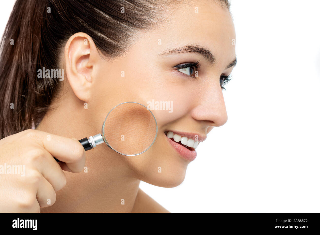 Close up conceptual portrait of young woman holding magnifying glass against cheek. Rough skin compared to smooth skin on face. Stock Photo