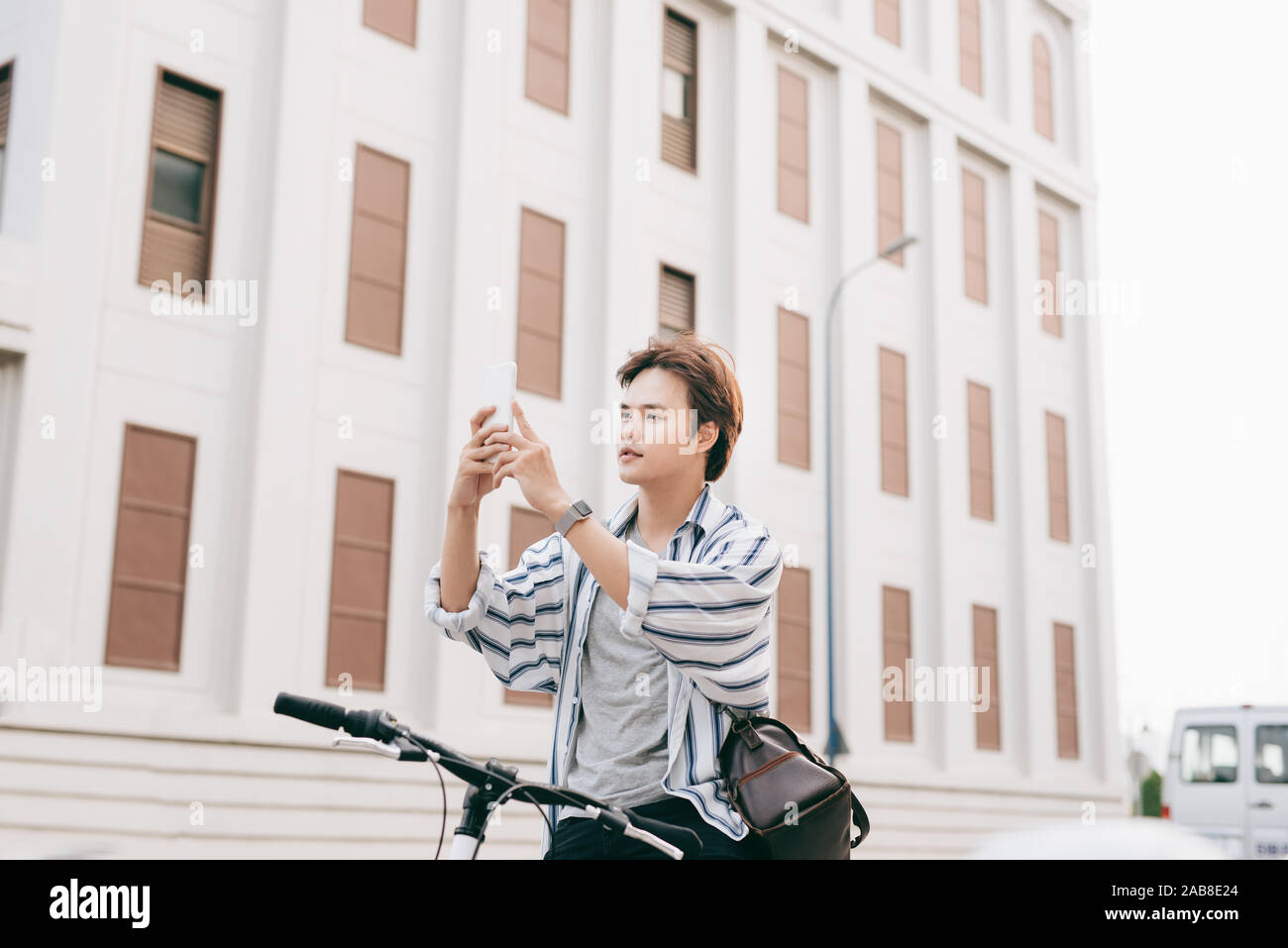 Stylish man chatting while using mobile phone while sitting on bicycle, outdoors. Dressed up in plaid shirt, t-shirt and jeans. Close-up. Stock Photo