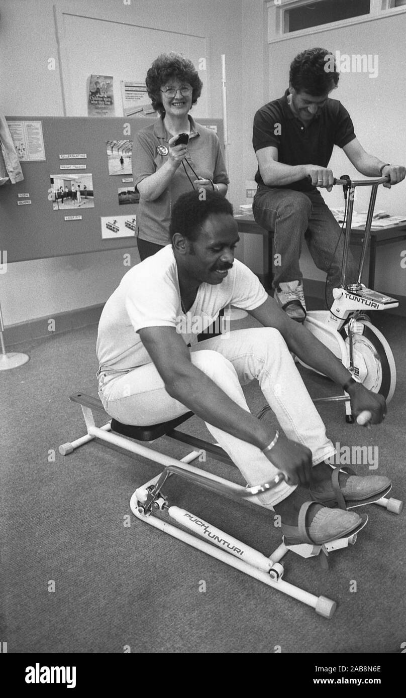 1980s, historical, healthly hearts, inside an office, staff using home gym kit, a rowing machine and an indoor exercise bike, inativive to improve mployees health, England, UK. Stock Photo