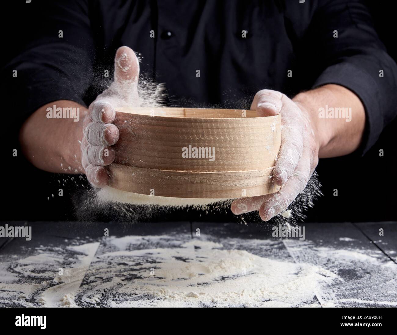 man sifts white wheat flour through a wooden sieve, black background ...