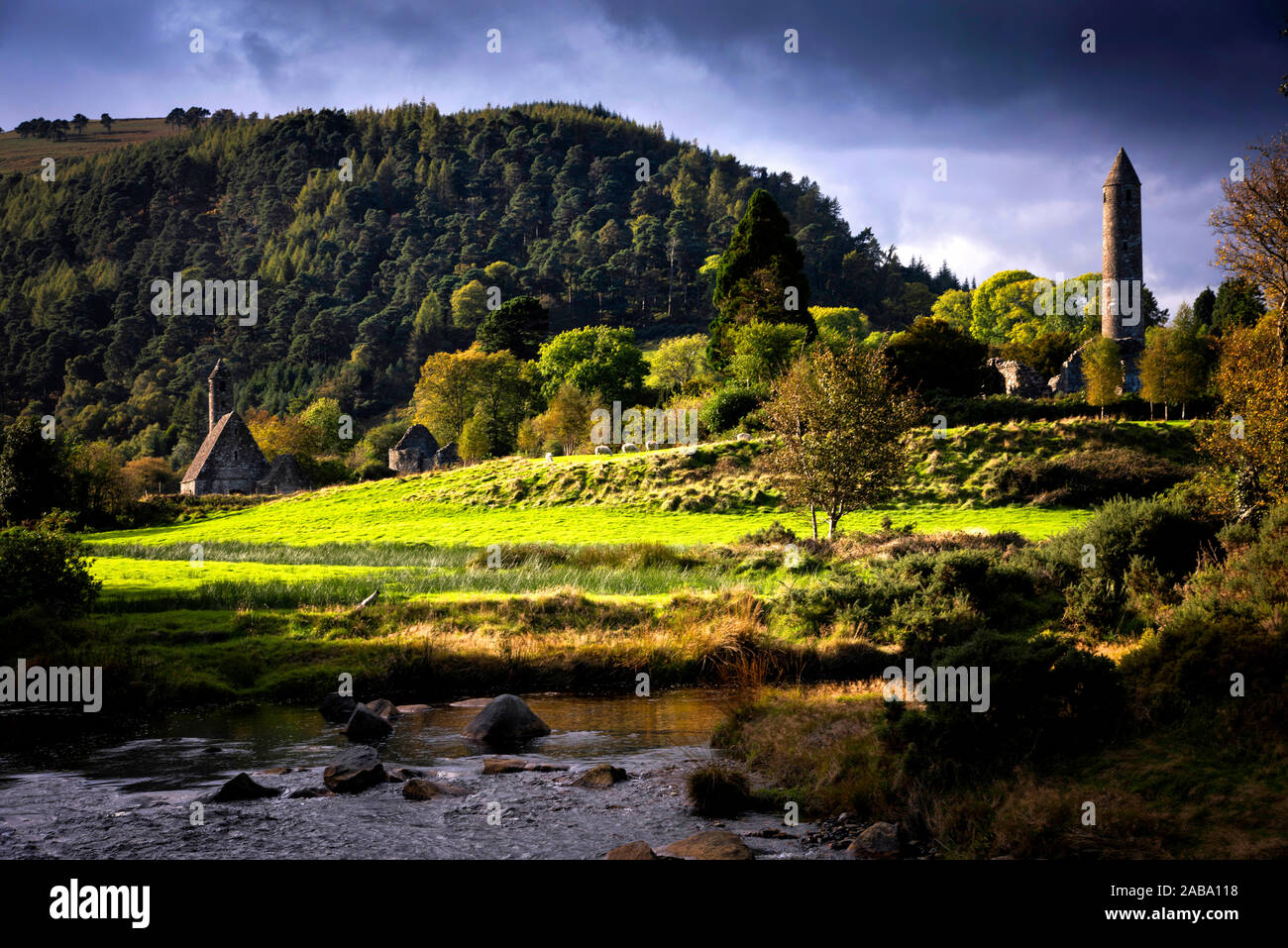 A river winds it's way past church and round tower at Glendalough Stock Photo