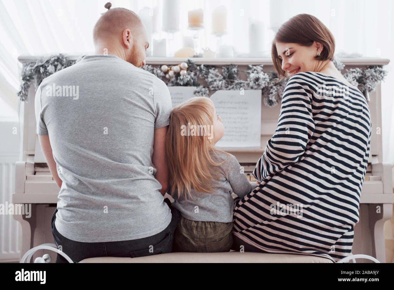 Look from behind at mother father and daughter playing white piano Stock Photo