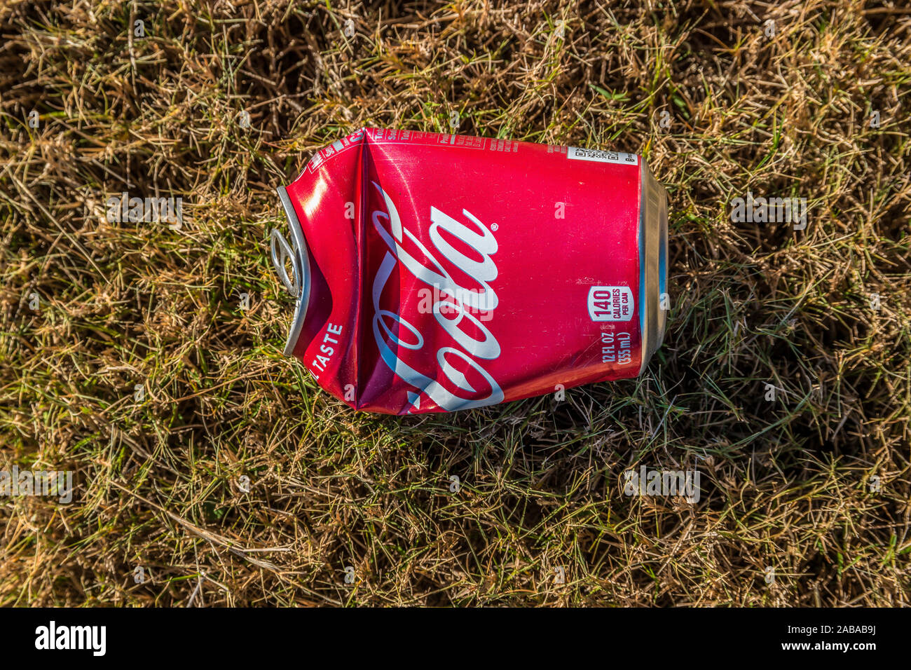 A horizontal view looking downward closeup at a bright red crushed Coca-Cola can laying on the grass outdoors on a sunny day in autumn Stock Photo