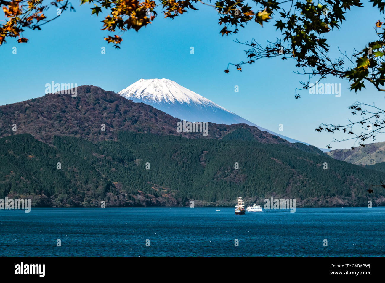 Mt. Fuji seen from Lake Ashinoko with two tourist cruise boats, shot through autumn foliage. Stock Photo