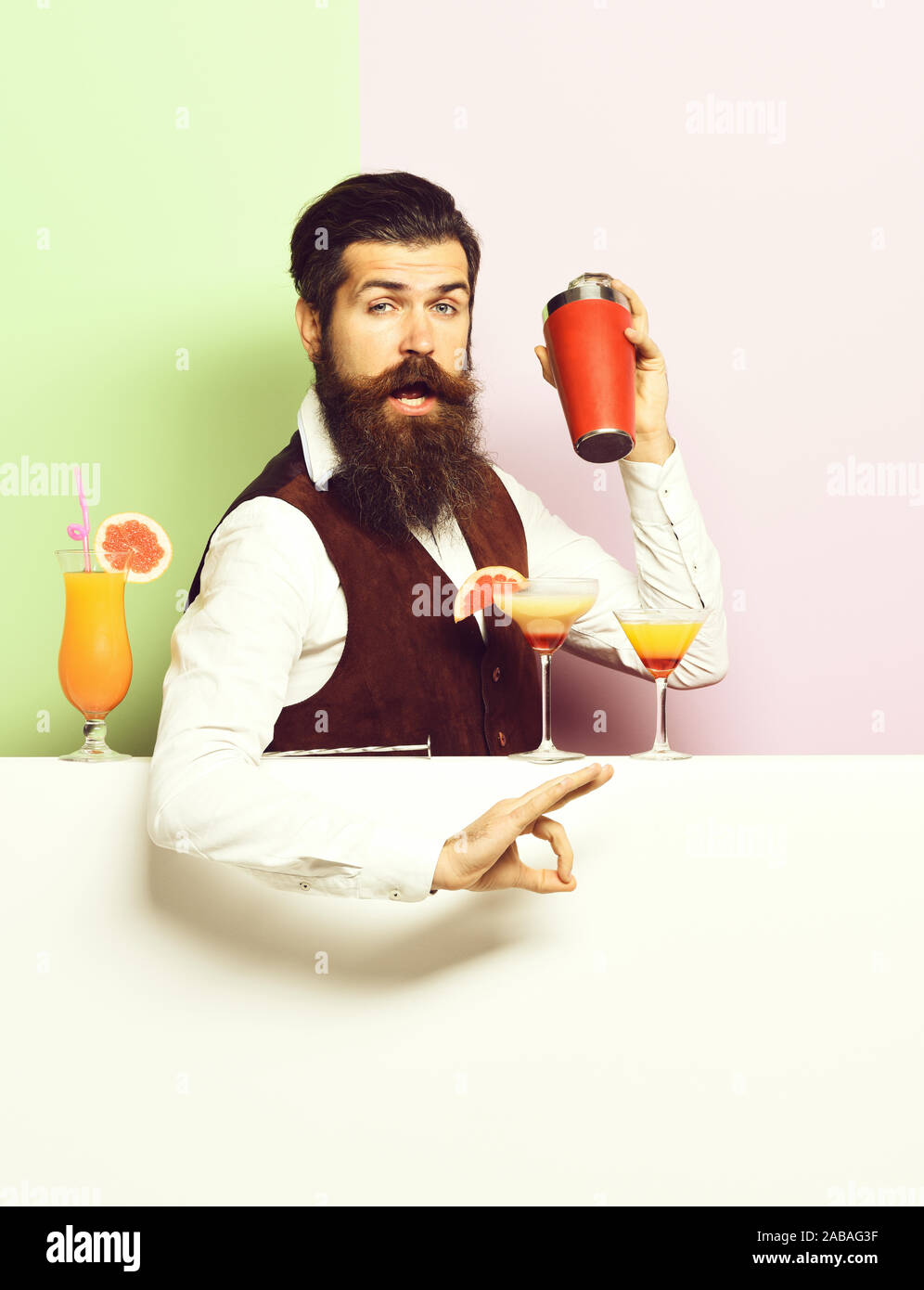 handsome bearded barman with long beard and mustache has stylish hair on funny face holding shaker. and made alcoholic cocktail in vintage suede leather waistcoat on purple green studio background Stock Photo