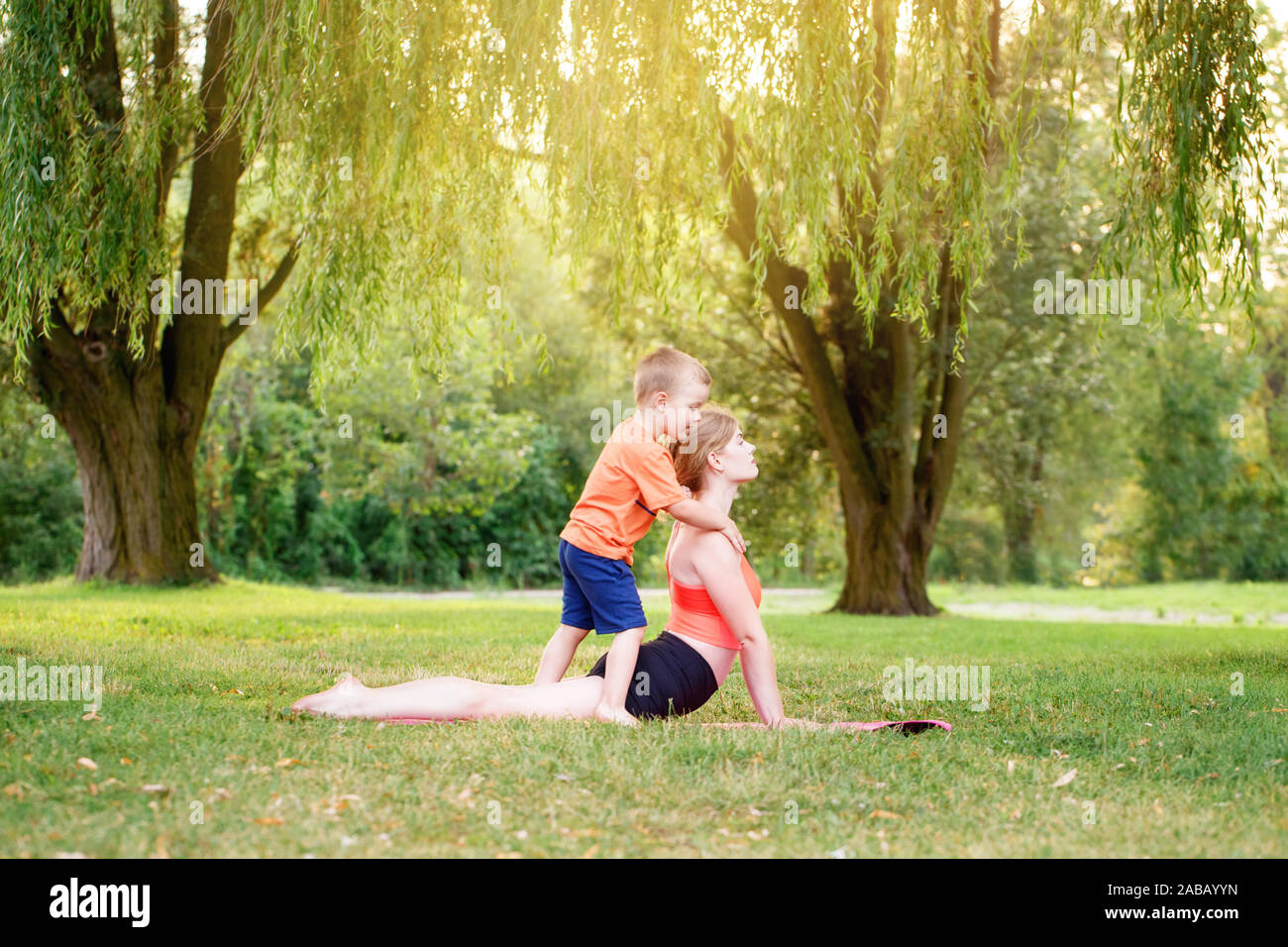 Family sport activity. Young Caucasian mother with child toddler boy doing workout yoga fitness outdoor in park on summer day. Woman doing exercises t Stock Photo