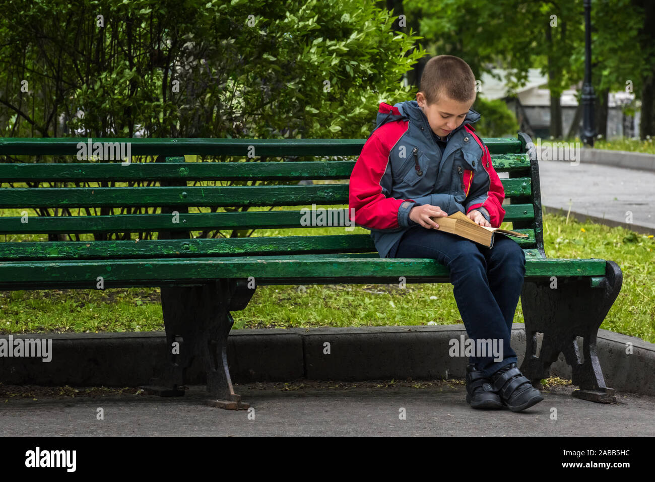Serious boy in casual clothing is reading a book. Boy is sitting alone on old painted green wooden bench in park. Selective focus. Unfocused green par Stock Photo