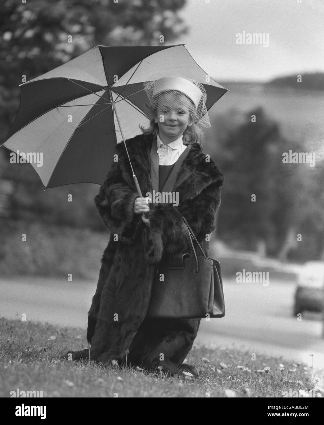 1980s, historical, outside holding an umbrella, a sweet little girl 'modelling' a previous generation's lady's clothing and accessories;  a thick fur coat, lace hat and handbag, all to be sold at a her school's jumble sale, England, UK. Stock Photo