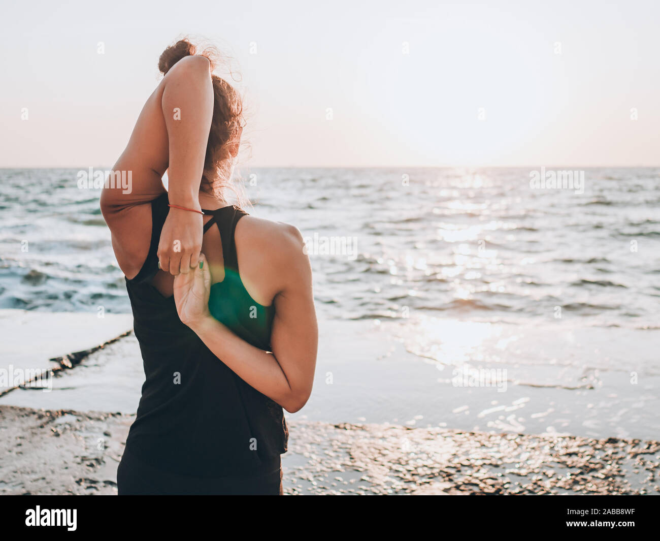 Slim woman in black bodysuit practicing yoga near sea or ocean during sunrise light. Flexibility, stretching, fitness, healthy lifestyle.  Stock Photo