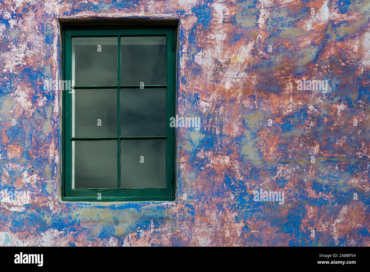 Window with green wood trim set in a weathered plaster wall with a distressed paint style in vibrant colors of blue, purple, and pink Stock Photo