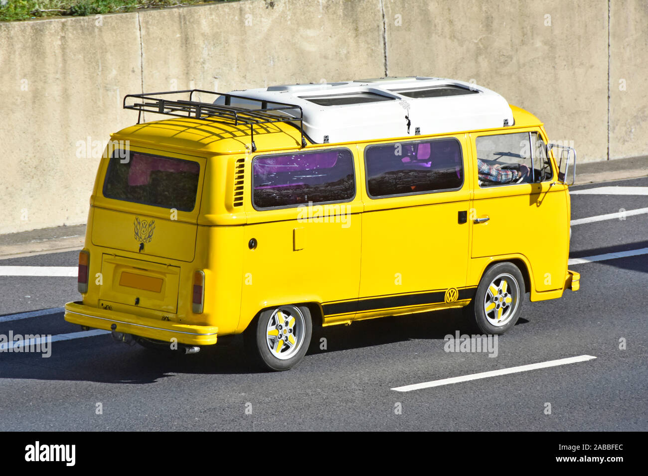 Side & back view of tinted windows on yellow VW Volkswagen camper van with white roof &  rear luggage rack driving along M25 motorway Essex England UK Stock Photo