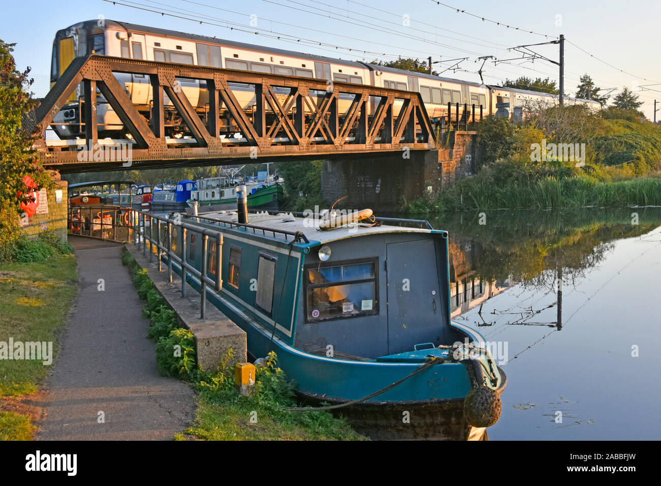 Stansted Express passenger train approaching Ely railway station on steel bridge above River Great Ouse narrowboat & towpath East Anglia England UK Stock Photo