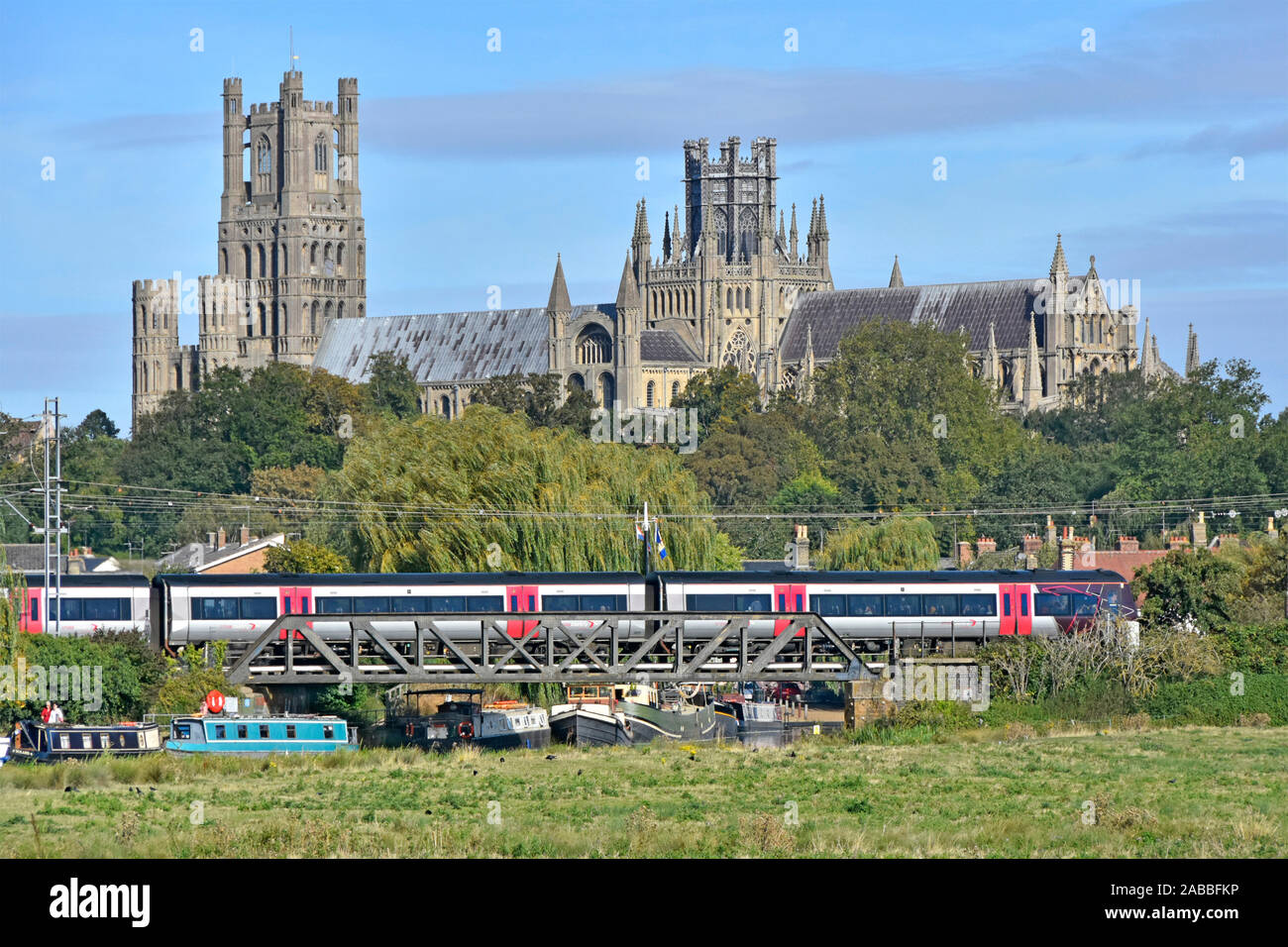 Ely Cathedral beyond train departing Ely Station Cambridgeshire boats on River Great Ouse below railway bridge  East Anglia Fens landscape England UK Stock Photo