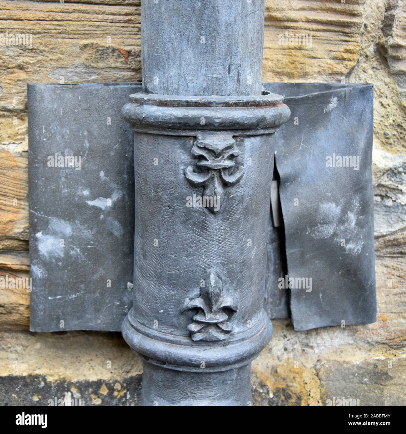 Close up 4” lead rainwater downpipe & fleur de lis ornamental design on plumbing straight joint held on wall with wide lead strap Durham cathedral UK Stock Photo