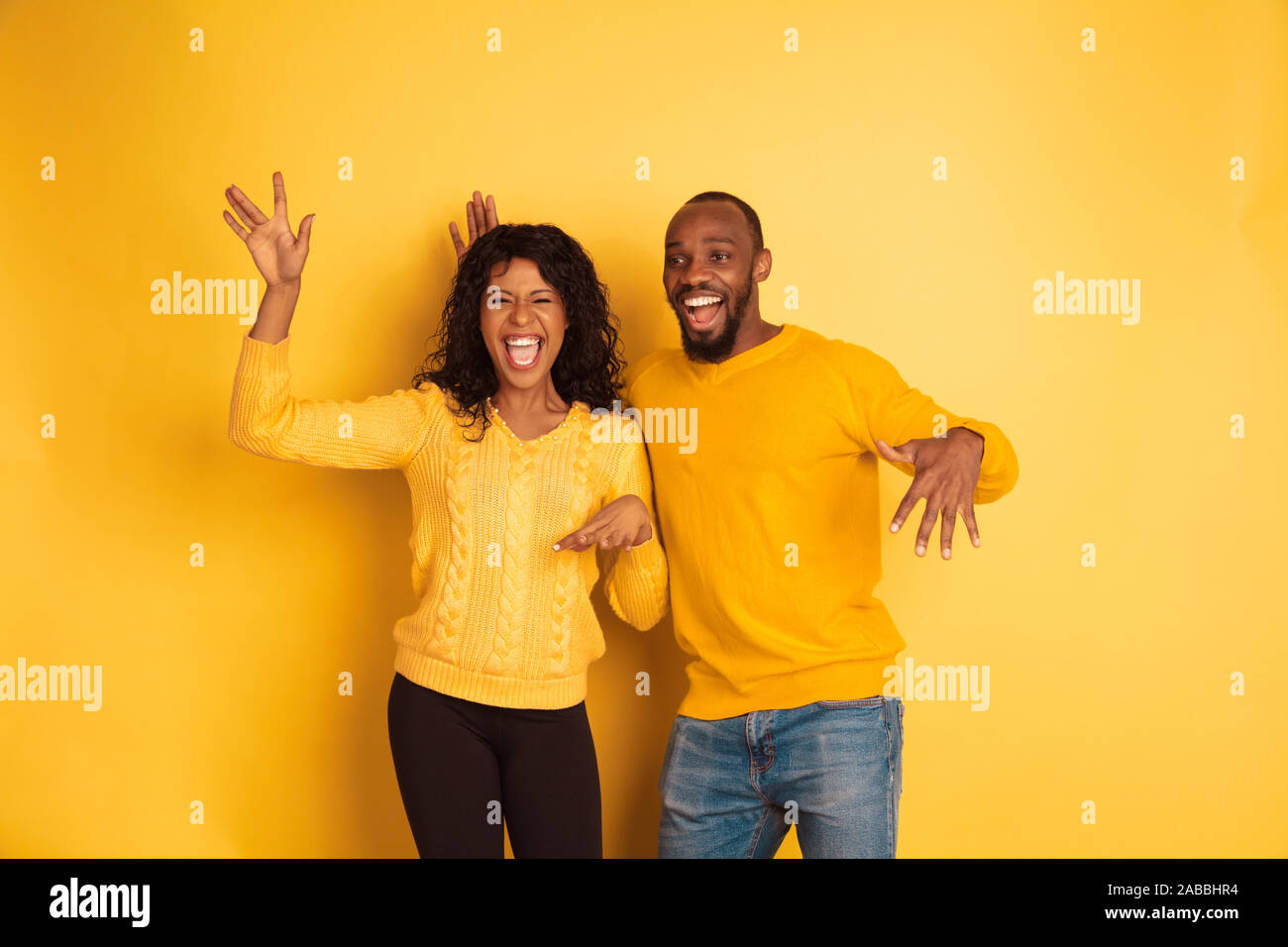Young emotional african-american man and woman in bright casual clothes on yellow background. Beautiful couple. Concept of human emotions, facial expession, relations, ad. Dancing and singing. Stock Photo